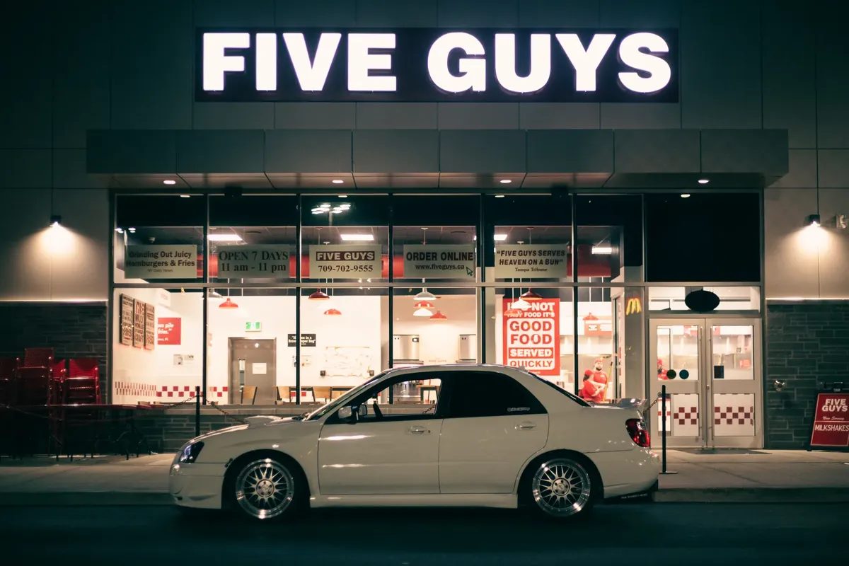 Illuminated Five Guys restaurant at night with a parked white Subaru Impreza.