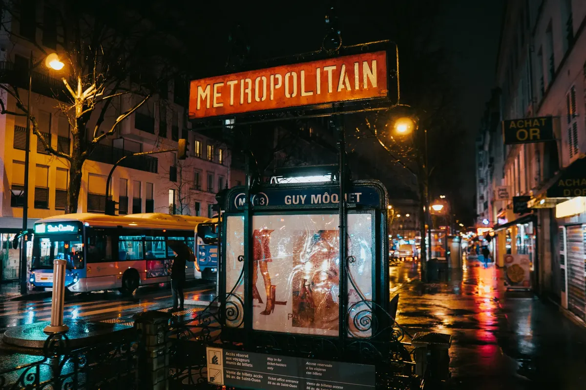 A wet Paris street captures the glow of city lights by the Metropolitain entrance at Guy Môquet metro station.